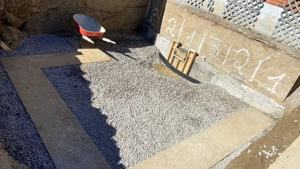 Subterranean Blueprint: Preparing the Groundwork A construction site with a gravel bed ready for foundation work, surrounded by concrete forms, with tools and a wheelbarrow