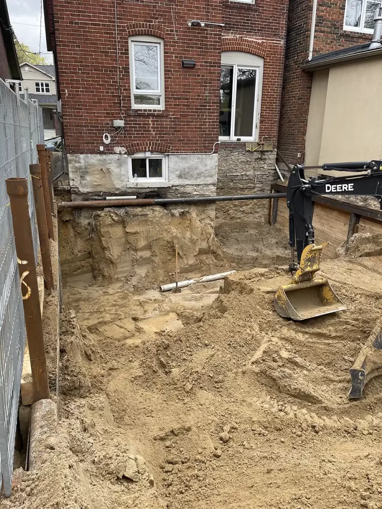 Initial Groundwork: Excavation for Structural Enhancement An excavator and a large dug-out pit beside a brick house, preparing for a construction project