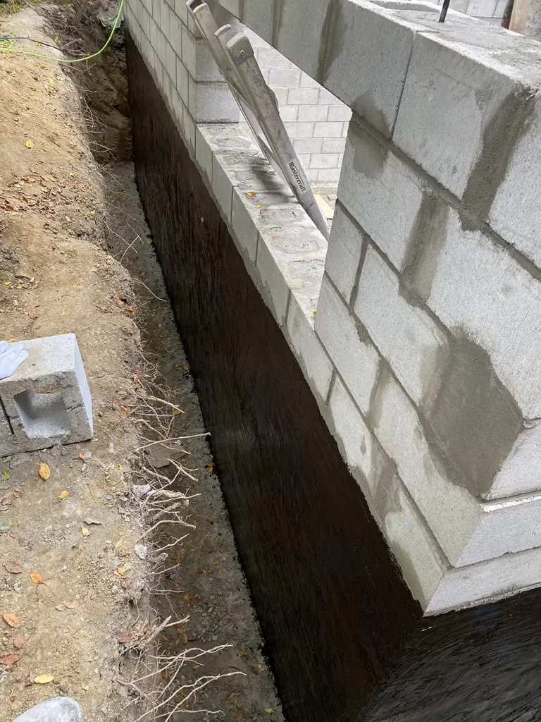 Sealed for Strength: Waterproofing in Construction A newly waterproofed foundation wall with a coating of black sealant, construction tools, and debris on the ground at a building site