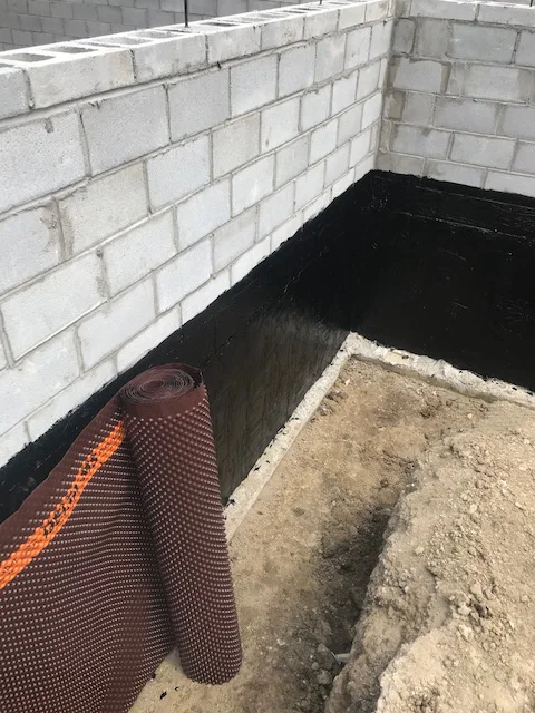 Waterproofing in Action: Securing the Foundation Part of a cinder block foundation wall coated in black waterproofing material, with a roll of waterproofing membrane nearby