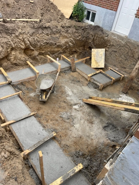 The Blueprint in Concrete: Foundation Footings Formed A construction site with newly poured concrete footings within wooden forms, a wheelbarrow, and excavation in the background.