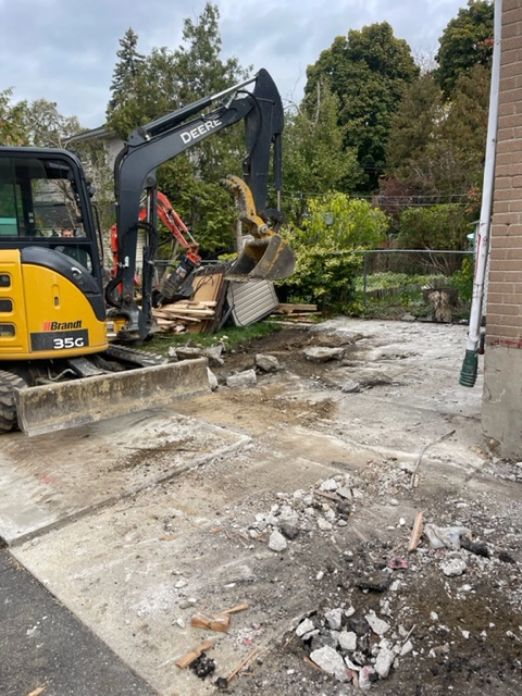A yellow excavator breaking up concrete in a residential construction area, preparing the site for new development.