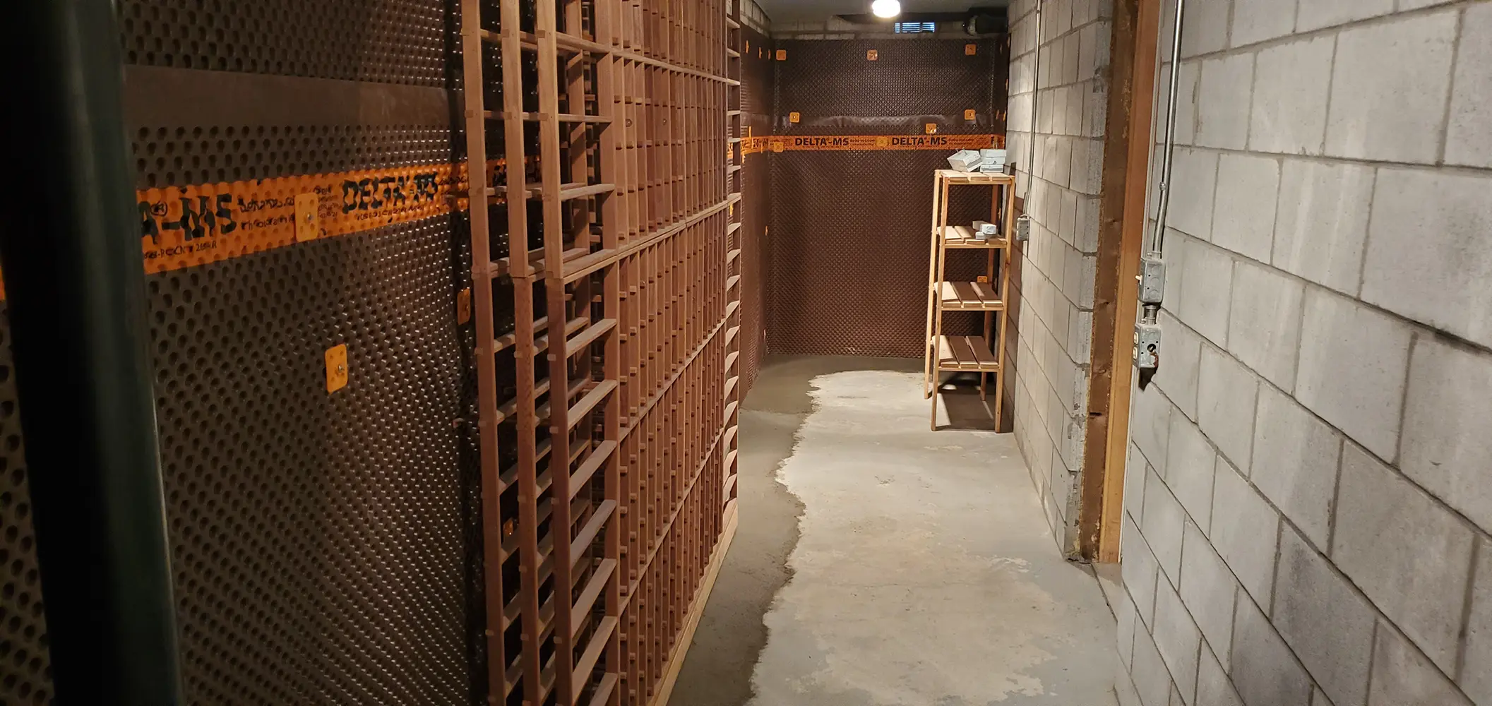 Interior view of a basement undergoing renovation with a waterproofing membrane on one wall, bare cinder blocks on the opposite wall, and a wooden shelving unit between them on a partially concreted floor.