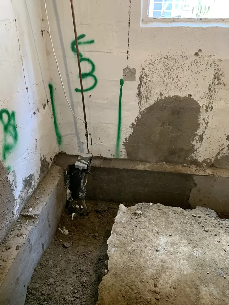 Initial Phases of Foundation Fortification Basement corner with exposed soil and partial concrete removal for foundation work, with green construction markings on the wall.