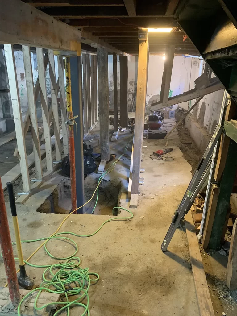 Framework of the Future: Basement Reinforcement Underway "A basement with a mix of wooden and metal supports, construction tools, and ongoing excavation work for reinforcement.