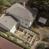 Captured from an aerial perspective, this image details an active construction site at the rear of a residential home. The foundations for an extension are clearly visible, with the wooden framework outlining the new addition. Construction equipment and materials are on site, and the presence of workers indicates ongoing development. The surrounding area includes neighboring houses, vehicles, and mature trees, providing context to the suburban setting.