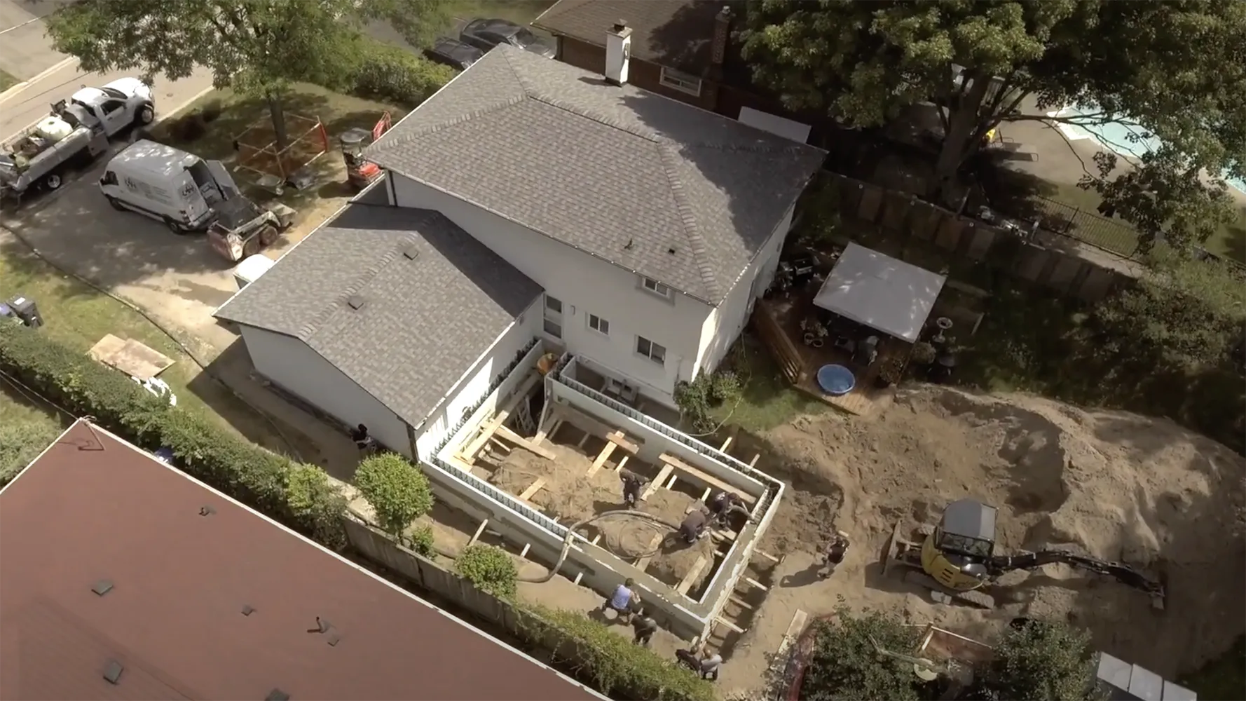 Captured from an aerial perspective, this image details an active construction site at the rear of a residential home. The foundations for an extension are clearly visible, with the wooden framework outlining the new addition. Construction equipment and materials are on site, and the presence of workers indicates ongoing development. The surrounding area includes neighboring houses, vehicles, and mature trees, providing context to the suburban setting.