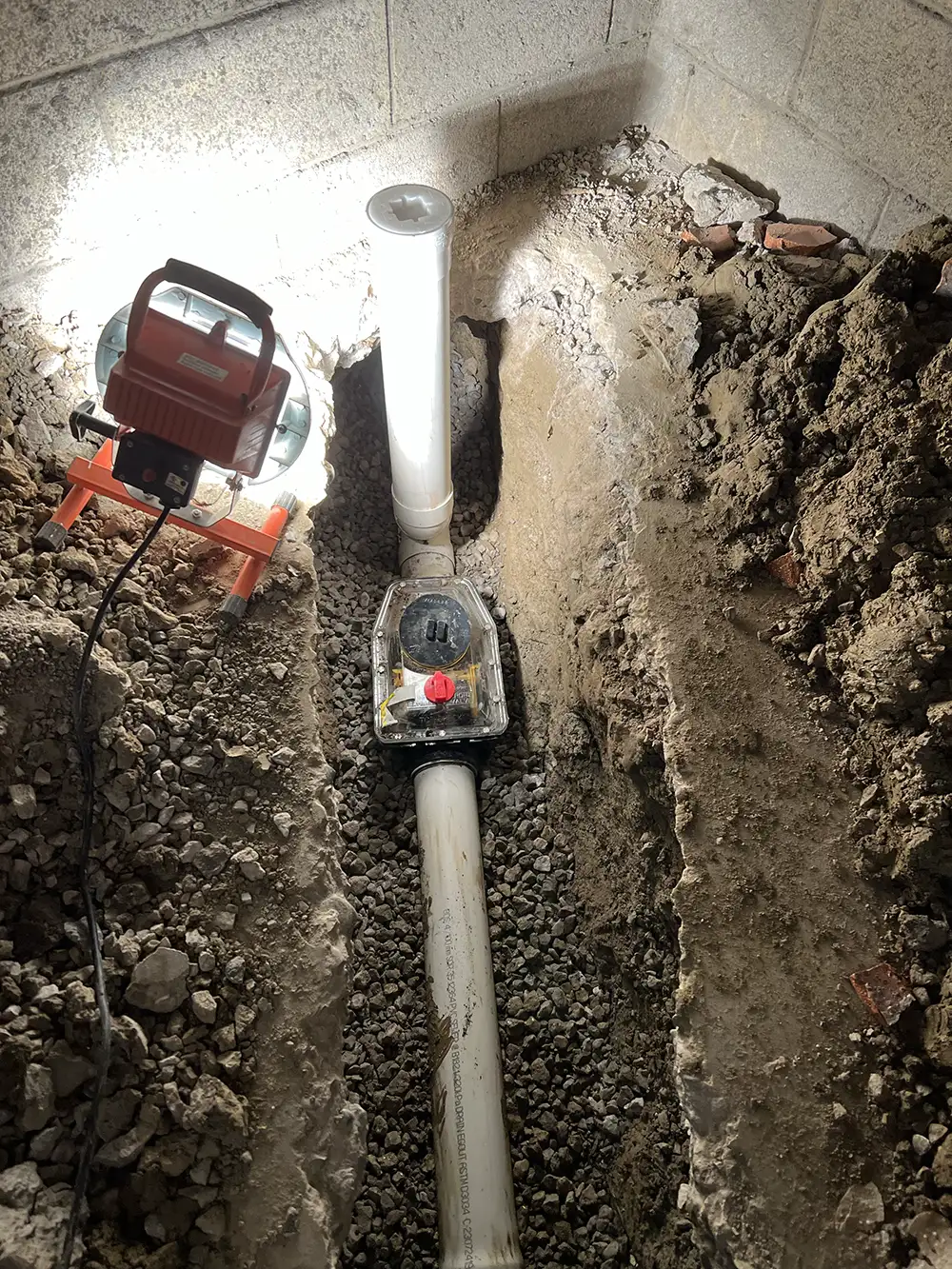 An excavated site for backwater valve installation with a new valve in place, lit by a portable work light, symbolizing proactive home protection