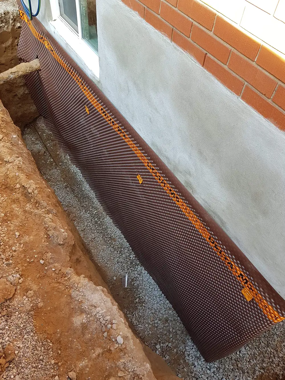 Brown waterproofing membrane with orange sealing tape installed along the exterior foundation of a house with a brick wall above and a trench filled with gravel below