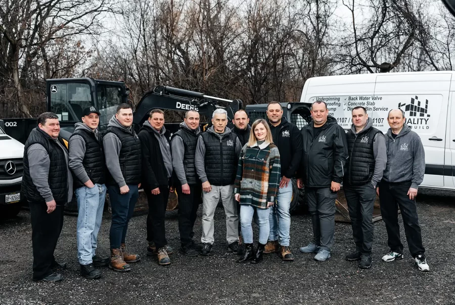 A group of eleven men and one woman from Quality Age Build, wearing coordinated workwear, stands before construction vehicles, showcasing their team spirit and professional dedication