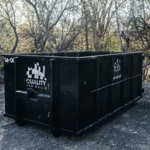 A 14-yard black garbage bin from Quality Age Build, placed on a gravel area with early spring trees in the background.