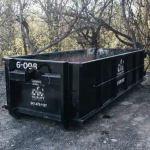 A 6-yard tall black garbage bin from Quality Age Build, positioned on a dusty ground with leafy trees in the backdrop.
