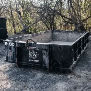 A wide 6-yard black garbage bin from Quality Age Build, resting on a dusty surface with a natural backdrop of trees.