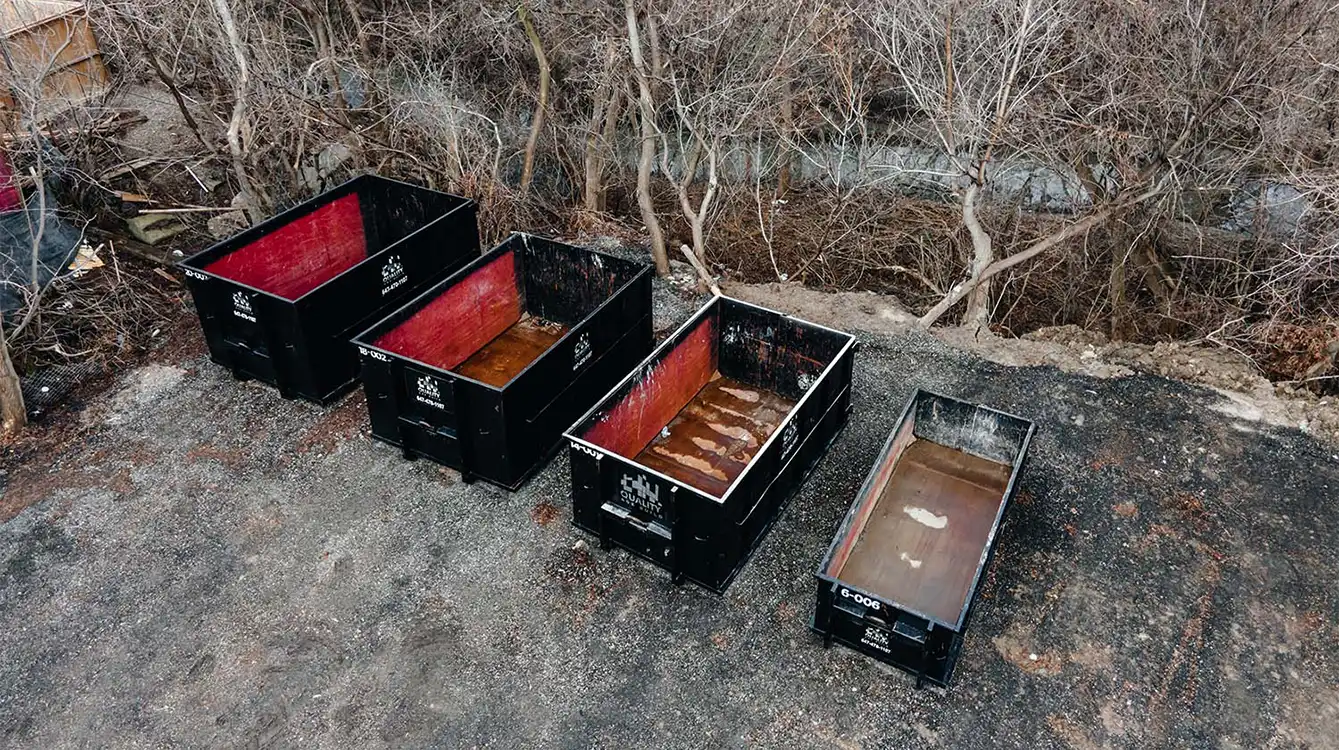 Aerial view of six large industrial garbage bins, varying in rust and usage wear, placed on a gravel ground surrounded by bare trees.