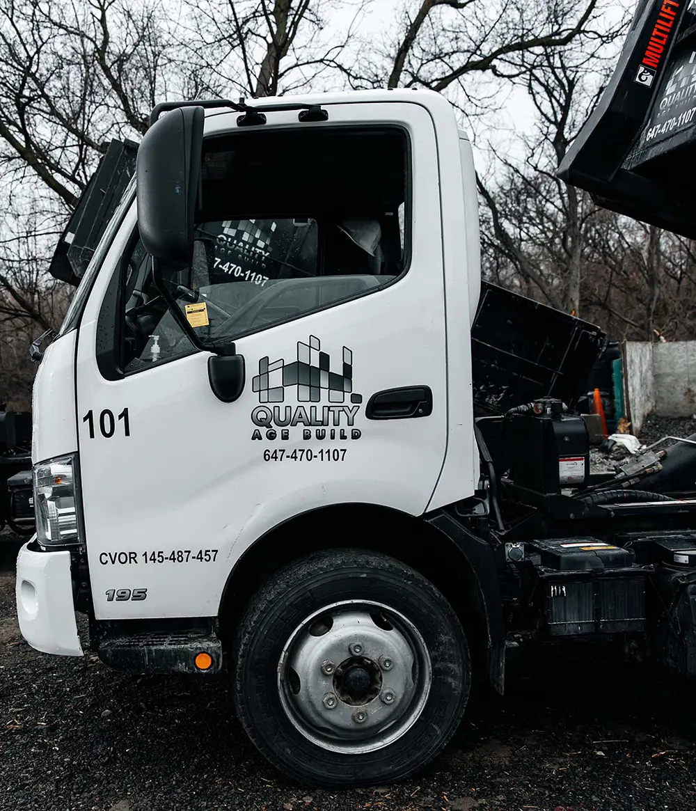 Close-up of a white Quality Age Build garbage bin truck, numbered 101, with an open bin lifting mechanism, against a backdrop of bare trees.