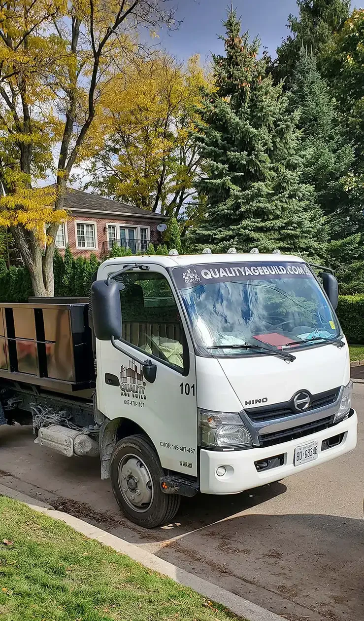 A white Quality Age Build garbage bin truck, numbered 101, parked on a residential street with autumn trees in the background.